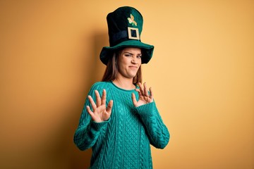 Young beautiful brunette woman wearing green hat with clover celebrating saint patricks day disgusted expression, displeased and fearful doing disgust face because aversion reaction. With hands raised