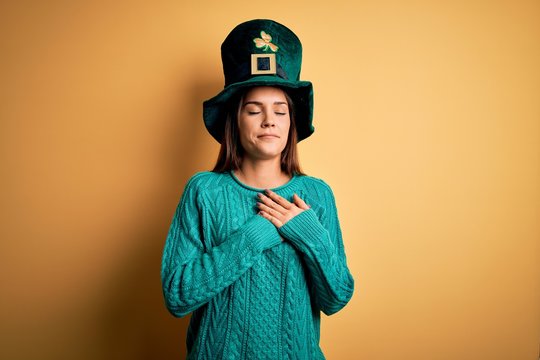 Young beautiful brunette woman wearing green hat with clover celebrating saint patricks day smiling with hands on chest with closed eyes and grateful gesture on face. Health concept.
