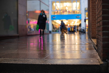 Out of focus picture of a woman shopping in the city centre with reflections of lights and store fronts on a blurry wet pavement - obrazy, fototapety, plakaty