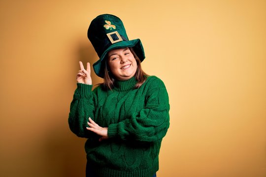 Young beautiful plus size woman wearing green hat with clover celebrating saint patricks day smiling with happy face winking at the camera doing victory sign. Number two.