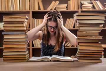 Young girl student reads a textbook sitting at a table with many books in the library