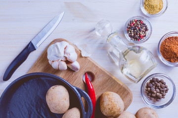 Potato, chili pepper, pan, knife, different spices and garlic on a white wooden background.