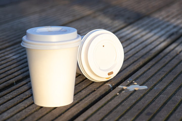 Closeup of two white single-use paper coffee cups with a blank surface on wooden background, outdoors in the morning