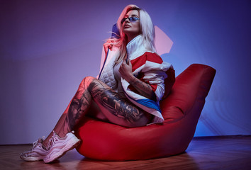 Attractive girl with tattooed leg is sitting on bean bag while posing for photographer.
