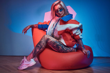 Attractive girl with tattooed leg is sitting on bean bag while posing for photographer.