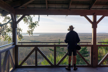 Fototapeta premium A senior man on a large veranda takes in the beautiful landscape background of a lowcountry salt marsh near Sapelo Island, coastal Georgia, USA.