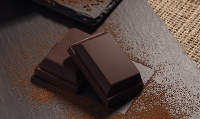 Two pieces of dark chocolate on black stone trays covered with cocoa powder top view