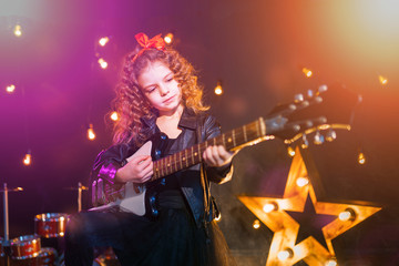Portrait of a Beautiful rock girl with curly hair wearing leather jacket, boots playing the electric guitar behind a red tank in recording studio.