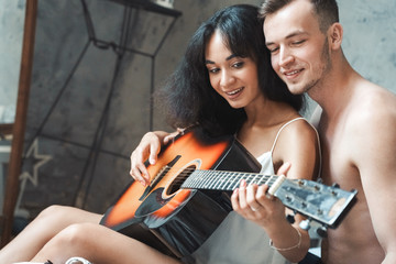 Fototapeta na wymiar Mixed Race Couple. Young man teaching woman playing guitar sitting on bed together smiling joyful