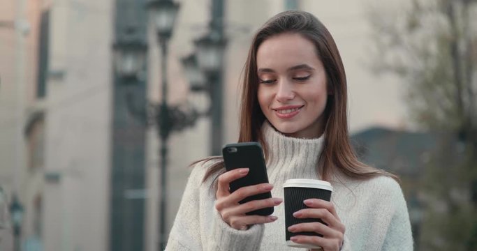 Portrait of Pretty Young Woman Standing in the City with Coffee to go and Mobile Phone. Wearing stylish Nude Sweater and having nice Makeup. Looking gorgeous and having charming Appearance
