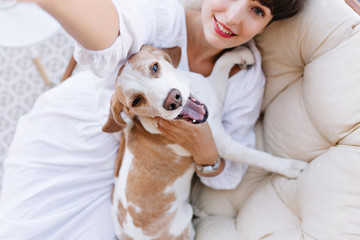 Girl with gray eyes and charming smile lies on soft sofa and making selfie with her pet. Indoor photo from above of brunette lady relaxing with funny beagle dog.