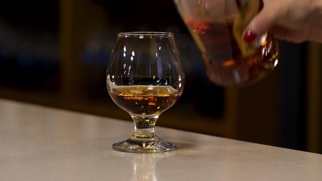 Close up of woman hand pouring cognac to the glass standing on the bar counter. Stock footage. Hand pouring alcohol into a glass on the blurred background of a dark room.