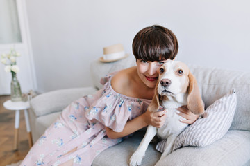 Brown-haired smiling girl in pink dress playing with her cute beagle dog at home. Indoor portrait of lovely young lady with dark short hair enjoying morning with pet, lying on sofa.