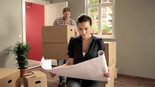 WS of a woman looking at blueprints while movers carry boxes in background