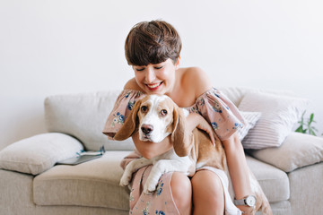 Pretty laughing female model holding beautiful shy puppy while posing on the couch. Indoor portrait of lovely girl with short shiny hair sitting on gray sofa with beagle dog and smiling.