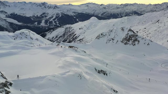 Drone shot tilting up to show ski fields with ski lifts and a frozen lake then sunset over a mountain horizon. In La Plagne France