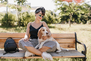Attractive brunette lady in striped pants sitting with legs crossed and stroking beagle dog....