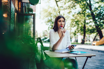Pensive hipster girl sitting at street cafeteria spending time for planning coursework organisation using public internet connection for research via cellphone device, pondering female student