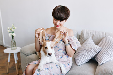 Amazing young woman wears striped pants and wristwatch posing on the floor while playing with beagle dog. Indoor portrait of happy girl with nude makeup spending time at home with puppy.