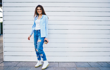 Full length portrait of attractive hipster girl in trendy outfit enjoying free time in city posing near copy space area for advertising,cheerful young woman in jeans apparel looking at camera outdoors