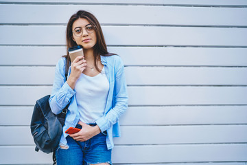 Half length portrait of charming beautiful woman in eyewear and stylish apparel posing on urban promotional background, young trendy dressed hipster girl holding mobile phone and coffee to go