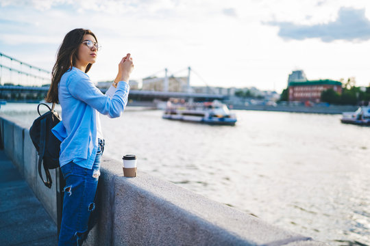 Young woman in casual wear holding mobile phone shooting video for travel vlog visiting city on sunny day, young millennial hipster girl influencer using smartphone for taking picture and share
