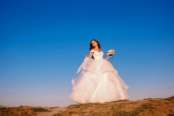 Stylish, elegant bride in a white dress stands on a mountain and poses on a background of blue sky, sunset