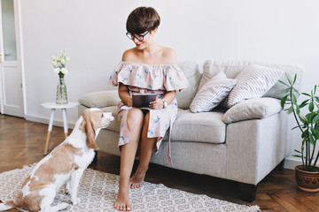 Barefooted smiling girl with white manicure looking with love at dog after work with tablet. Indoor...