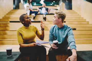 Diverse couple of learners discussing lecture while drinking coffee sitting at auditorium