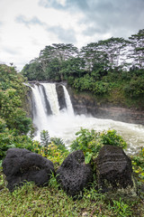 Hilo, Hawaii, USA. - January 14, 2020: White Rainbow Falls on foaming violent Wailuku River surrounded by green trees and plants under white-gray cloudscape.