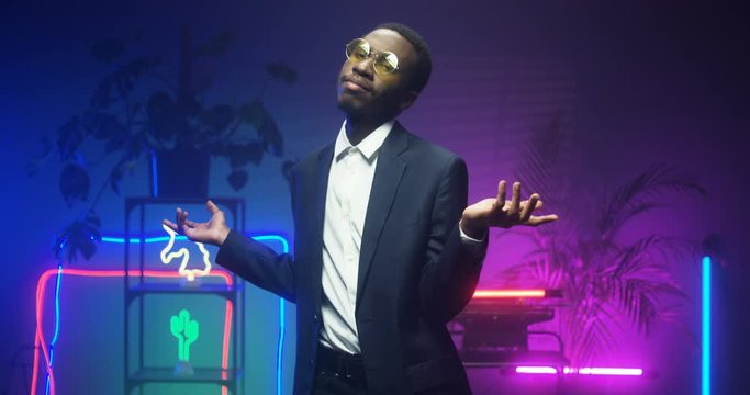 African american young handsome man in glasses and fancy suit dancing in room with neon lights. Black gentleman at futuristic party. Guy in LED pink lights. Male dancer.