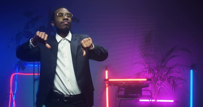 African American young handsome man in glasses and fancy suit giving his thumb down dancing in room with neon lights. Black gentleman at futuristic party. Guy in LED pink lights.
