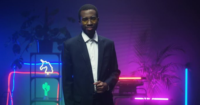 Young African American handsome gentleman in glasses and suit standing in room with blue and pink lights of neon tubes. Portrait of good looking man turning face to camera and smiling. Indoor.