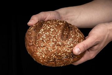 Man's hands hold tasty fresh loaf of dark bread with sesame seeds on black background