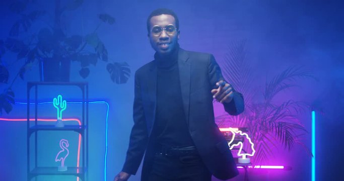 African American young handsome man in glasses dancing in room with neon lights. Black good looking male dancer at futuristic party alone. Guy in blue and pink lights of asteras.