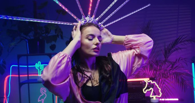 Portrait of beautiful young brunette Caucasian woman in crown from neon sticks with colorful lights in futuristic room. Close up of fantastic female beauty queen. Model striking a pose. Indoors.