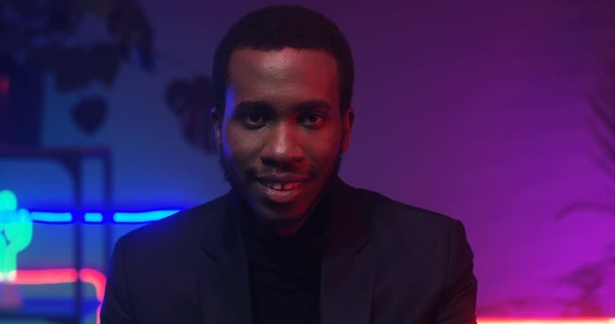Close up of African American young handsome man smiling to camera in room with neon pink and blue lights. Portrait of black guy in club atmosphere. Male model smile.