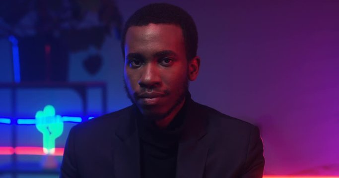 Close up of African American young handsome man looking to camera in room with neon pink and blue lights. Portrait of black guy in club atmosphere. Male model with serious face.