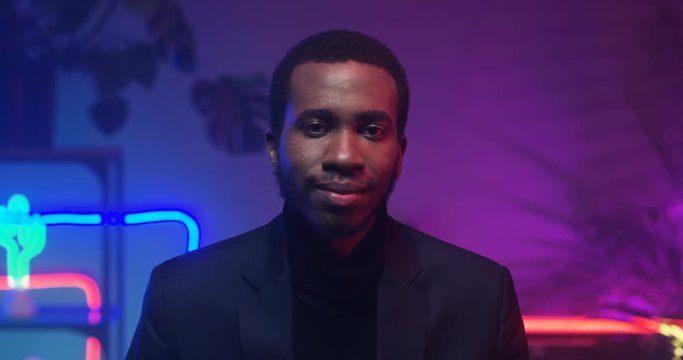 Close up of African American young attractive man looking to camera in room with neon pink and blue lights. Portrait of black guy in club atmosphere. Male model smile. Indoors.