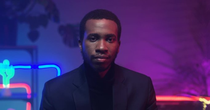 Blurred African American young handsome man looking to camera in room with neon blue and pink lights. Portrait of black male model. Camera zooming in on guy face. Indoors.