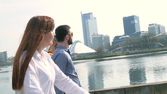 Young beautiful woman and three men are going along the city embankment with high-rise buildings in the background on sunny spring day in slow motion middle shot 4K video