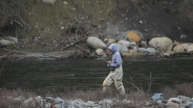 Male in waders alone fishing peacefully casting lure walking across on river water bank on sandy river in pacific northwest oregon wide angle in slow motion