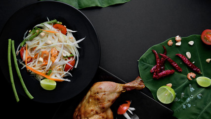 Top view of papaya salad, chicken grill on black plate and ingredients on green taro