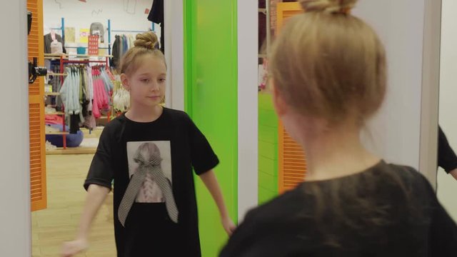 Adorable girl in stylish dress looking at mirror in fitting room. Beautiful teenage girl wearing stylish clothes and enjoying her reflection in mirror during shopping in mall