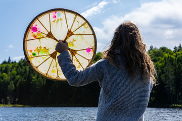 Rear view of young shamanic woman holding sacred native frame drum while in lake praying an...