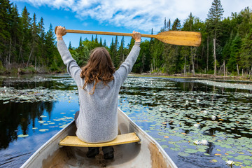 Rear view of young tourist holding paddle raised high while sitting in canoe riding in lake with...