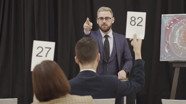 Elegant male auctioneer standing at lectern, pointing at paddles and announcing bids while selling contemporary painting at art auction