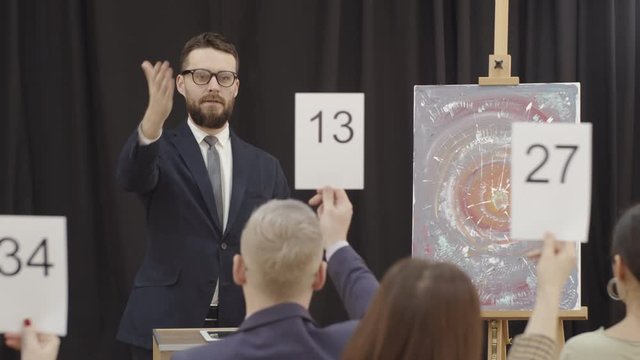 Bearded male auctioneer in formal suit pointing at paddles and calling out bids while standing at lectern in front of audience and selling modern artwork