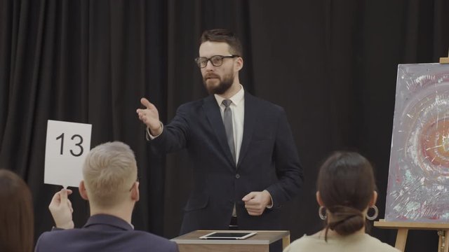 Male auctioneer pointing at paddle and calling bid while standing in front of participants and selling contemporary painting at art auction