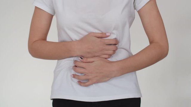 Close-up of women's hands clutching their sick stomach. The girl has a stomach cramp, she holds back the pain with her hands. Close-up of a sick woman's stomach in a white t-shirt on a white